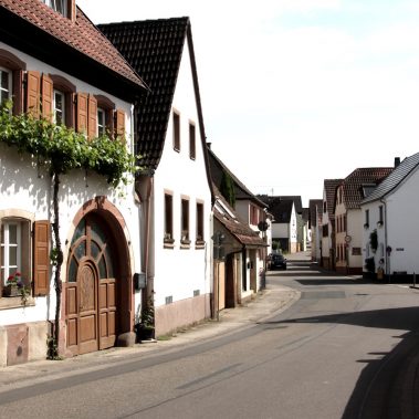 Eine idyllische Dorfstraße in Böchingen mit weißen historischen Fachwerkhäusern, braunen Fensterläden und einem Torbogen an der Südlichen Weinstraße.