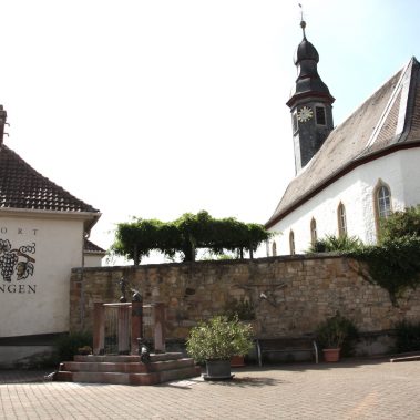 Die weiße Kirche von Böchingen mit markantem Kirchturm hinter einer alten Steinmauer, dem Dorfbrunnen und einer Hauswand mit der Aufschrift „Weinort Böchingen“.