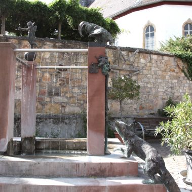 Der historische Dorfbrunnen aus Sandstein mit Tierfiguren vor einer alten Steinmauer und der Kirche in Böchingen.