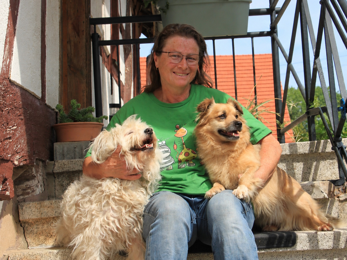 Doris Dühr-Bien sitzt lächelnd auf den Steinstufen am Eingang des Ferienhauses QuarTIER in Böchingen und hält zwei Hunde im Arm.