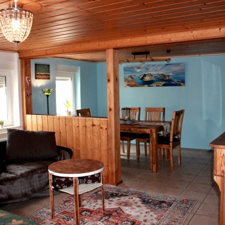 Ein gemütlicher Wohn- und Essbereich mit einer Couch, einem kleinen Couchtisch auf einem Orientteppich, einer halbhohen Holztrennwand und einem dahinterliegenden hölzernen Esstisch im Ferienhaus QuarTIER in Böchingen.